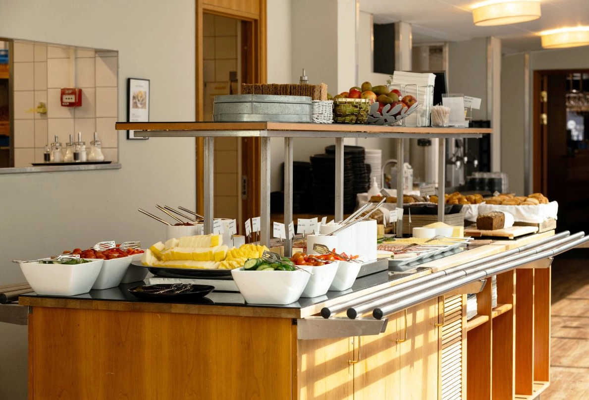 Frukostbuffébord i trä på Kom Hotel. Bordet är överfyllt av en uppdukad frukostbuffé med grönsaker, frukt och pålägg till smörgåsar.