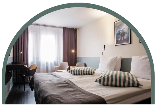 Bilden visar ett dubbelrum standard på Kom Hotel i Stockholm. Rummet har ett fönster inramat av gardiner, en dubbelsäng, skrivbord och två fåtöljer. Rummet har ett trägolv och väggarna pryds av två tavlor.