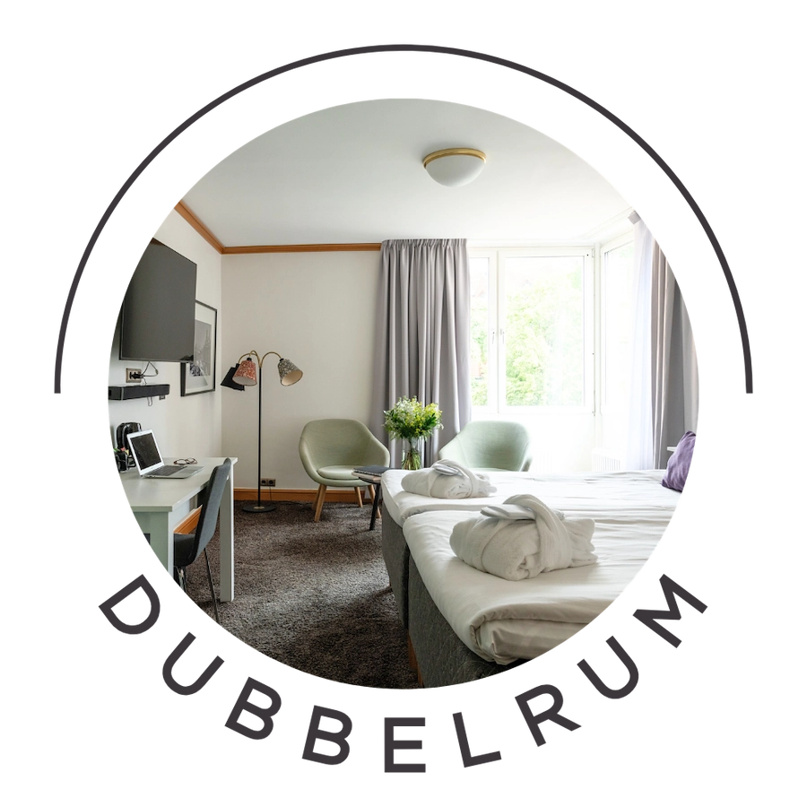 Bilden visar ett dubbelrum standard på Kom Hotel i Stockholm. Rummet har ett fönster inramat av gardiner, en dubbelsäng, skrivbord och två fåtöljer. Rummet har heltäckningsmatta och väggarna pryds av en TV.