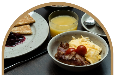 Uppdukad hotellfrukost på ett bord. Bacon med äggrora och tomat, pannkakor med sylt. Ett glas juice står på bordet.