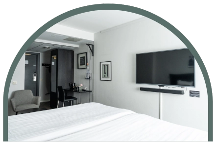 Bilden visar ett dubbelrum standard på Kom Hotel i Stockholm. Rummet har en dubbelsäng, en fåtölj, skrivbord, skrivbordsstol och en TV.  Det hänger två tavlor på de vita väggarna.