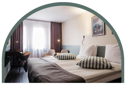Bilden visar ett dubbelrum standard på Kom Hotel i Stockholm. Rummet har ett fönster inramat av gardiner, en dubbelsäng, skrivbord och två fåtöljer. Rummet har ett trägolv och väggarna pryds av två tavlor.