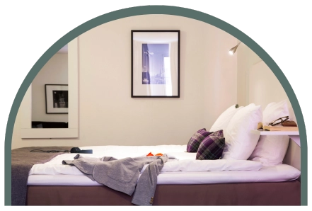 Bilden visar ett dubbelrum standard på Kom Hotel i Stockholm. Rummet har en dubbelsäng, en spegel, en tavla och gärgerna går i vitt uppblandat med jordnära nyanser.