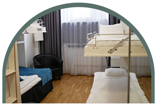Bilden visar ett ekonomirum cabin på Kom Hotel i Stockholm. Rummet har trägolv och ett fönster, 2 våningssängar med totalt 4 bäddar och en fåtölj. Fönstret ramas in av gardiner. 