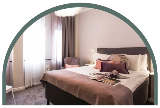 Bilden visar ett dubbelrum storlek small på Kom Hotel i Stockholm. Rummet har ett fönster, en dubbelsäng och en fåtölj med ett litet bord. Fönstret ramas in av gardiner och hotellrummets golv täcks av en heltäckningsmatta som går i samma färger som rummet.