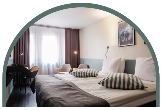 Bilden visar ett dubbelrum standard på Kom Hotel i Stockholm. Rummet har ett fönster inramat av gardiner, en dubbelsäng, skrivbord och två fåtöljer. Rummet har ett trägolv och väggarna pryds av två tavlor.
