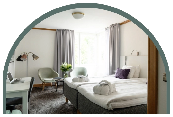 Bilden visar ett dubbelrum superior på Kom Hotel i Stockholm. Rummet har ett fönster inramat av gardiner, en dubbelsäng med två morgonrockar liggandes på sängen, skrivbord, två fåtöljer och ett bord. Rummet har en heltäckningsmatta och en golvlampa.