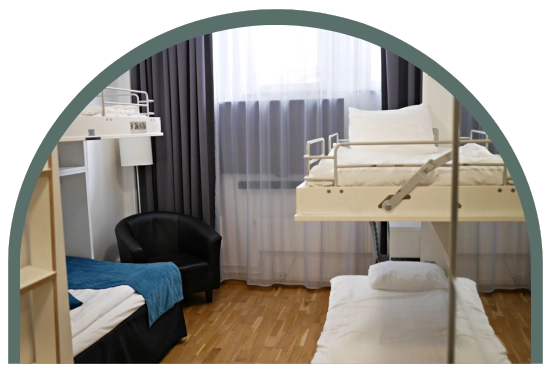 Bilden visar ett ekonomirum cabin på Kom Hotel i Stockholm. Rummet har trägolv och ett fönster, 2 våningssängar med totalt 4 bäddar och en fåtölj. Fönstret ramas in av gardiner. 
