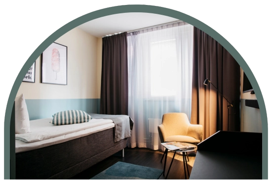 Bilden visar ett enkelrum på Kom Hotel. Rummet har ett fönster, en enkelsäng, skrivbord och en fåtölj med ett litet bord. Det sitter en tv på väggen och fönstret ramas in av gardiner. På väggen sitter två tavlor. Väggarna går i neutrala färger. 