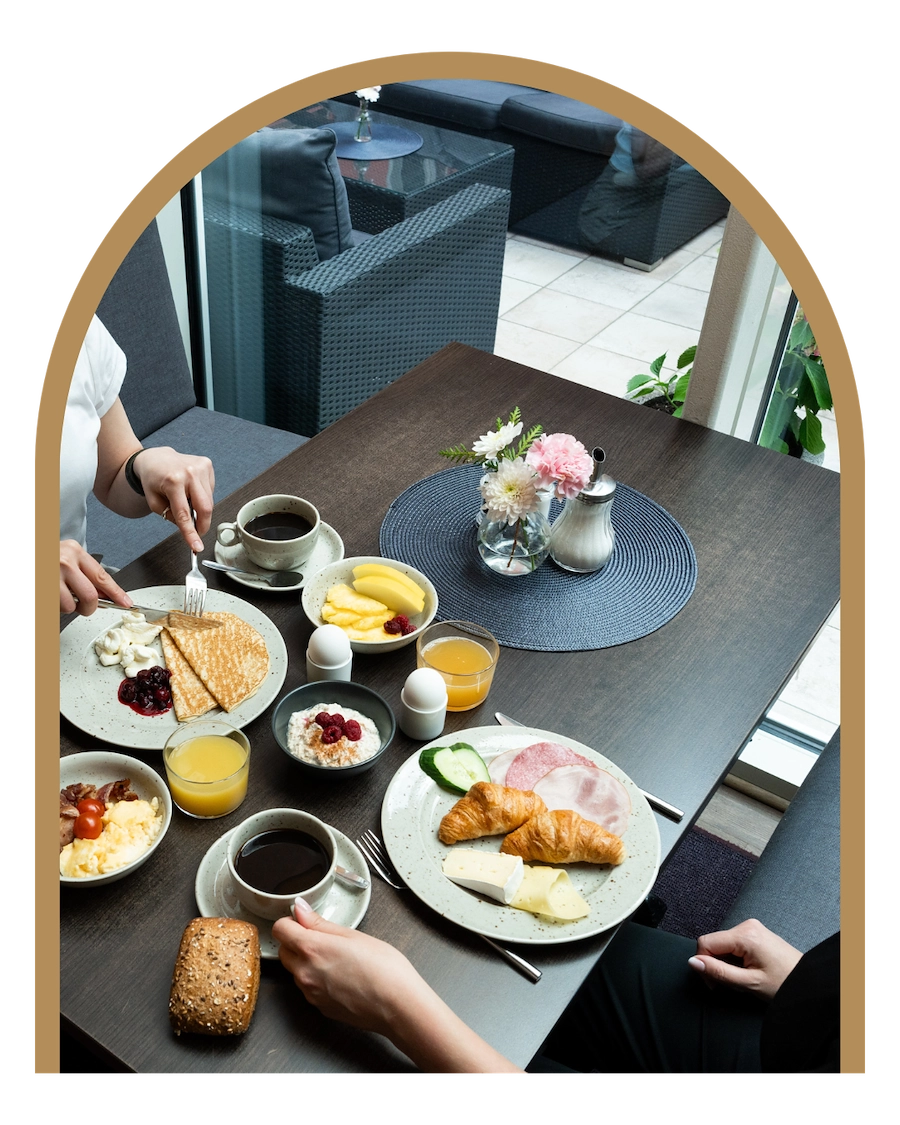 Bilden visar två personer som äter frukost. Bara bordet och dess händer syns i bild och de har bordet uppdukat med en härlig frukost innehållandes croissanter, kaffe, frukt, pannkakor och andra goda frukosttillbehör.