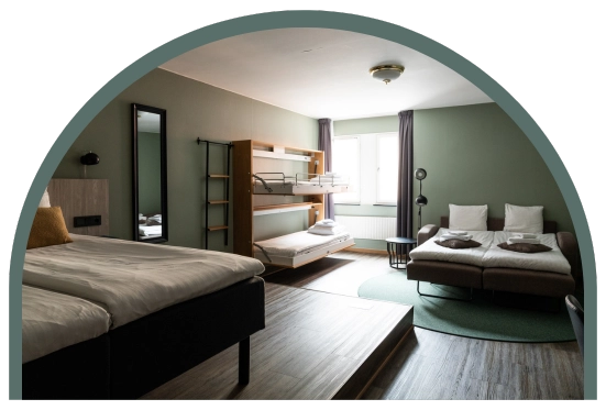 Bilden visar ett familjerum large på Kom Hotel i Stockholm. Rummet har ett fönster inramade av gardiner, en dubbelsäng, en bäddsoffa, en våningssäng, spegel och ett litet bord. Rummet har trägolv.