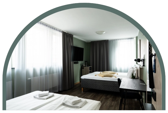 Bilden visar ett familjerum på Kom Hotel i Stockholm. Rummet har två fönster inramade av gardiner, en dubbelsäng, en bäddsoffa, skrivbord och en TV. Rummet har trägolv.