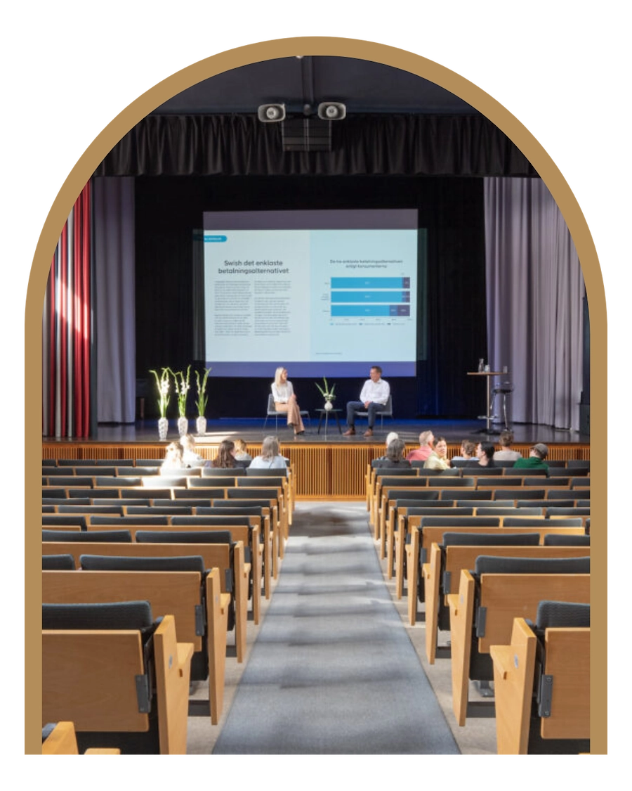 En stor konferenssal med mycket sittplatser i form av bänskar som vätter mot en stor scen. Några människor sitter i publiken och två människkor är på scenen.