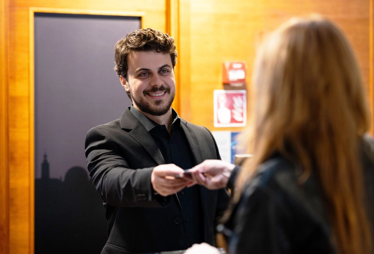 En manlig receptionist på Kom Hotel lämnar över en rumsnyckel till en gäst med ett leende på läpparna.