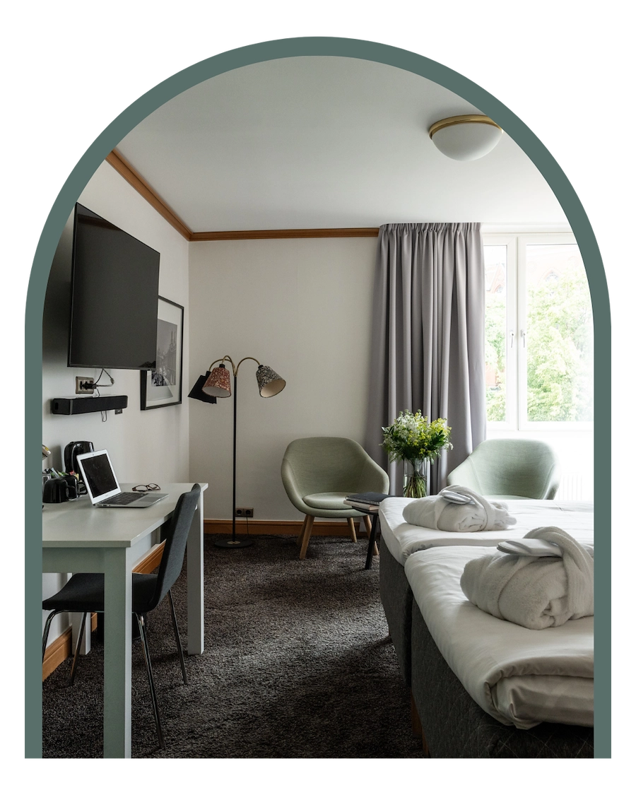 Bilden visar ett dubbelrum superior på Kom Hotel i Stockholm. Rummet har ett fönster inramat av gardiner, en dubbelsäng med två morgonrockar liggandes på sängen, skrivbord, två fåtöljer och ett bord. Rummet har en heltäckningsmatta och en fin golvlampa.