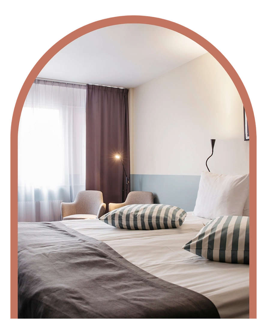 Bilden visar ett dubbelrum standard på Kom Hotel i Stockholm. Rummet har ett fönster inramat av gardiner, en dubbelsäng, skrivbord och två fåtöljer. Rummet har ett trägolv och väggarna pryds av två tavlor.