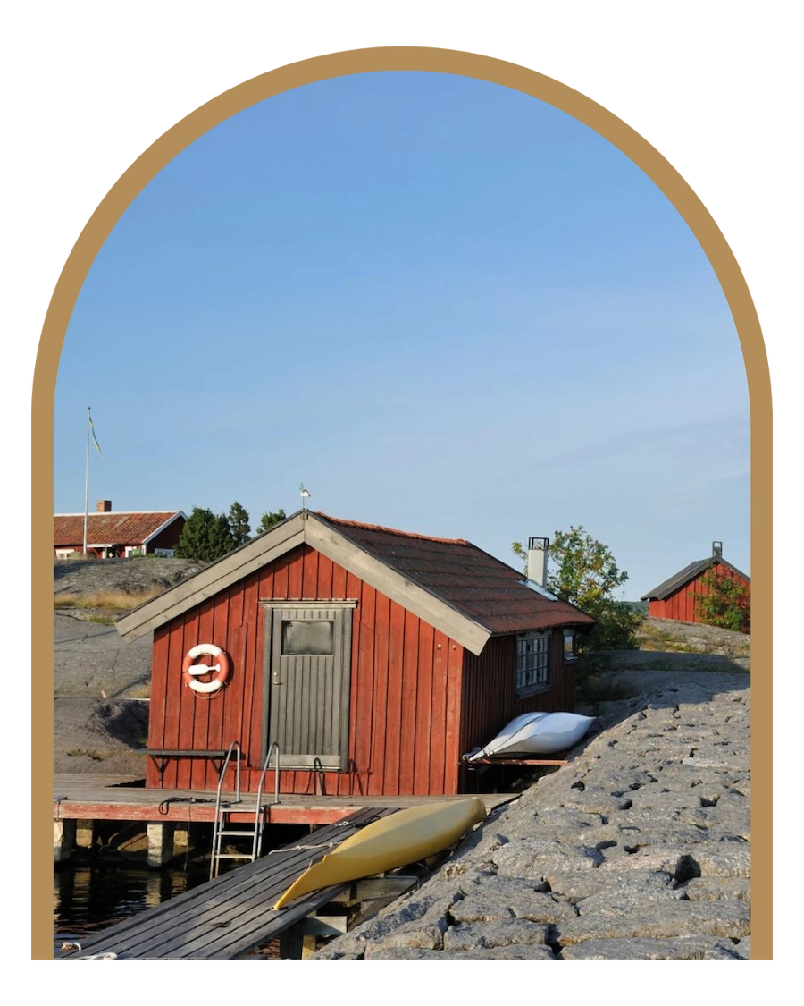 En typisk bild av en röd stuga i Stockholm Skärgård. Huset ligger på en berg och solar sig i solen. På väggen hänger en livboj i klassiskt röda och vita färger.