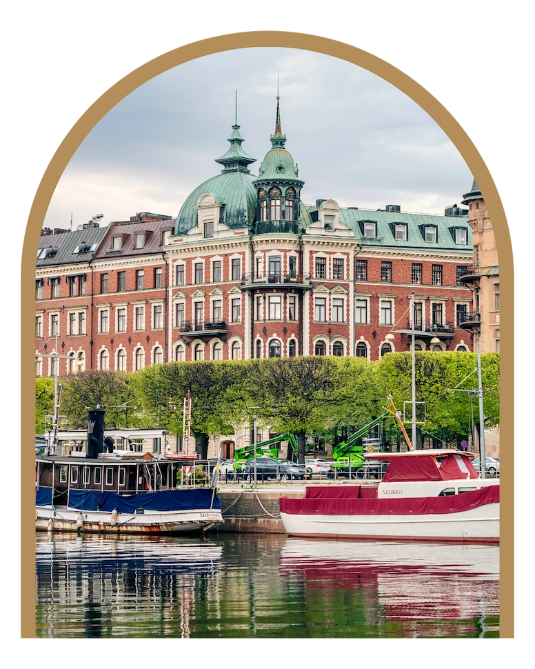 En bild på byggnaderna på Strandvägen i Stockholm sett från vattnet och Djurgården. I förgrunden ligger några husbåtar. Staden grönskar.