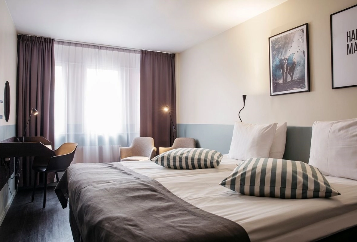 Bilden visar ett dubbelrum standard på Kom Hotel i Stockholm. Rummet har ett fönster inramat av gardiner, en dubbelsäng, skrivbord och två fåtöljer. Rummet har trägolv och väggarna pryds av en TV.